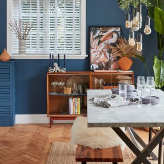 A blue-painted dining room with a marble-effect dining table and a sideboard displaying glassware