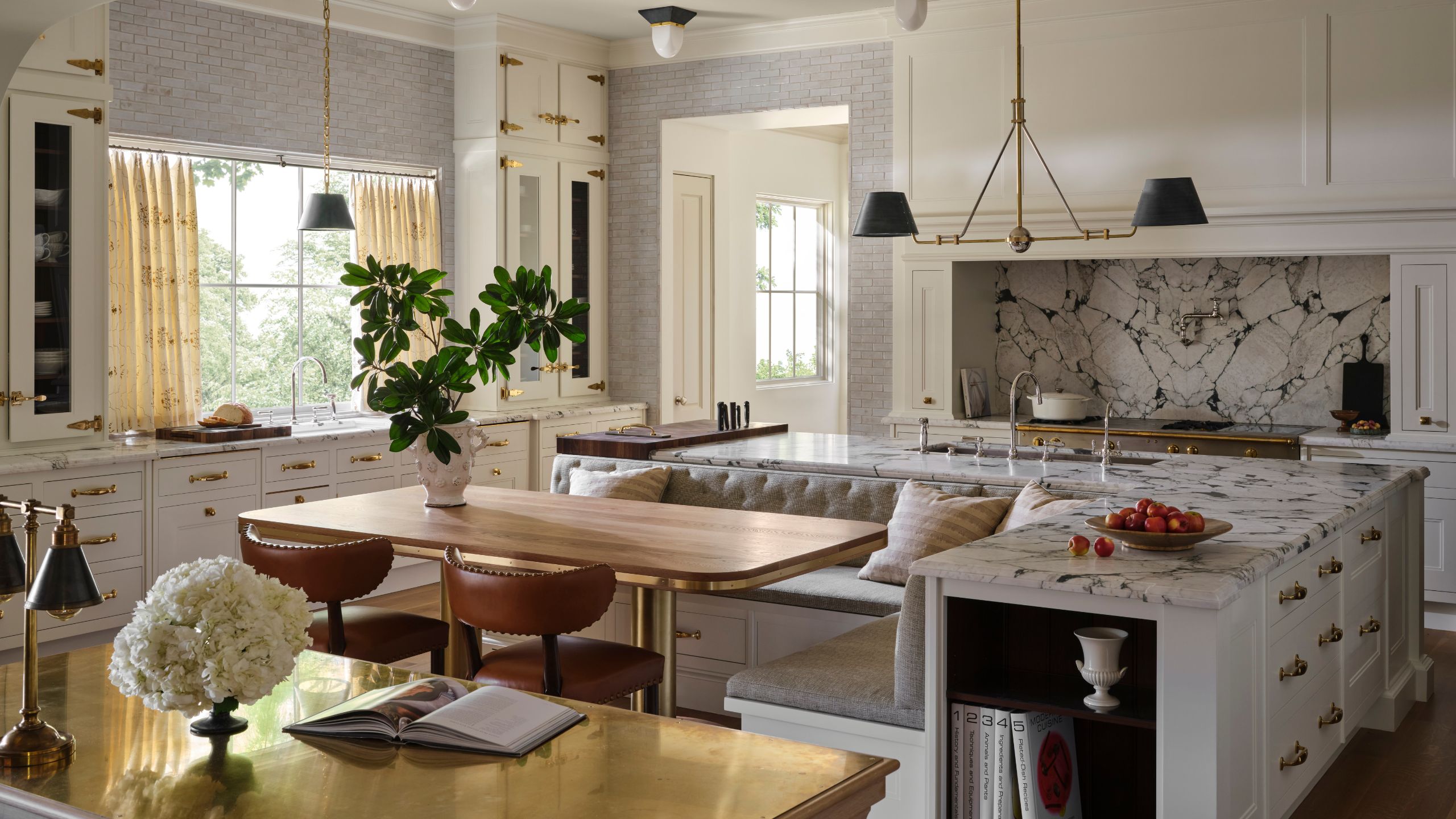 a warm soft white grand kitchen with banquette built into the island, marble backsplash and counters and layered lighting