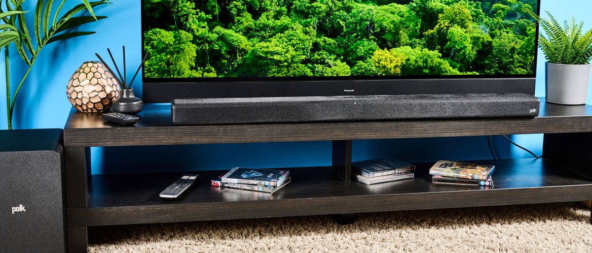 The Polk Audio Signa S4 being used in a lounge with a large TV. On the TV screen is a green forest scene. In the background is a blue wall.