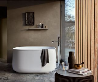 white soaking style freestanding bath in bathroom with tiled floor, wooden slatted screen and dark brown plaster effect walls