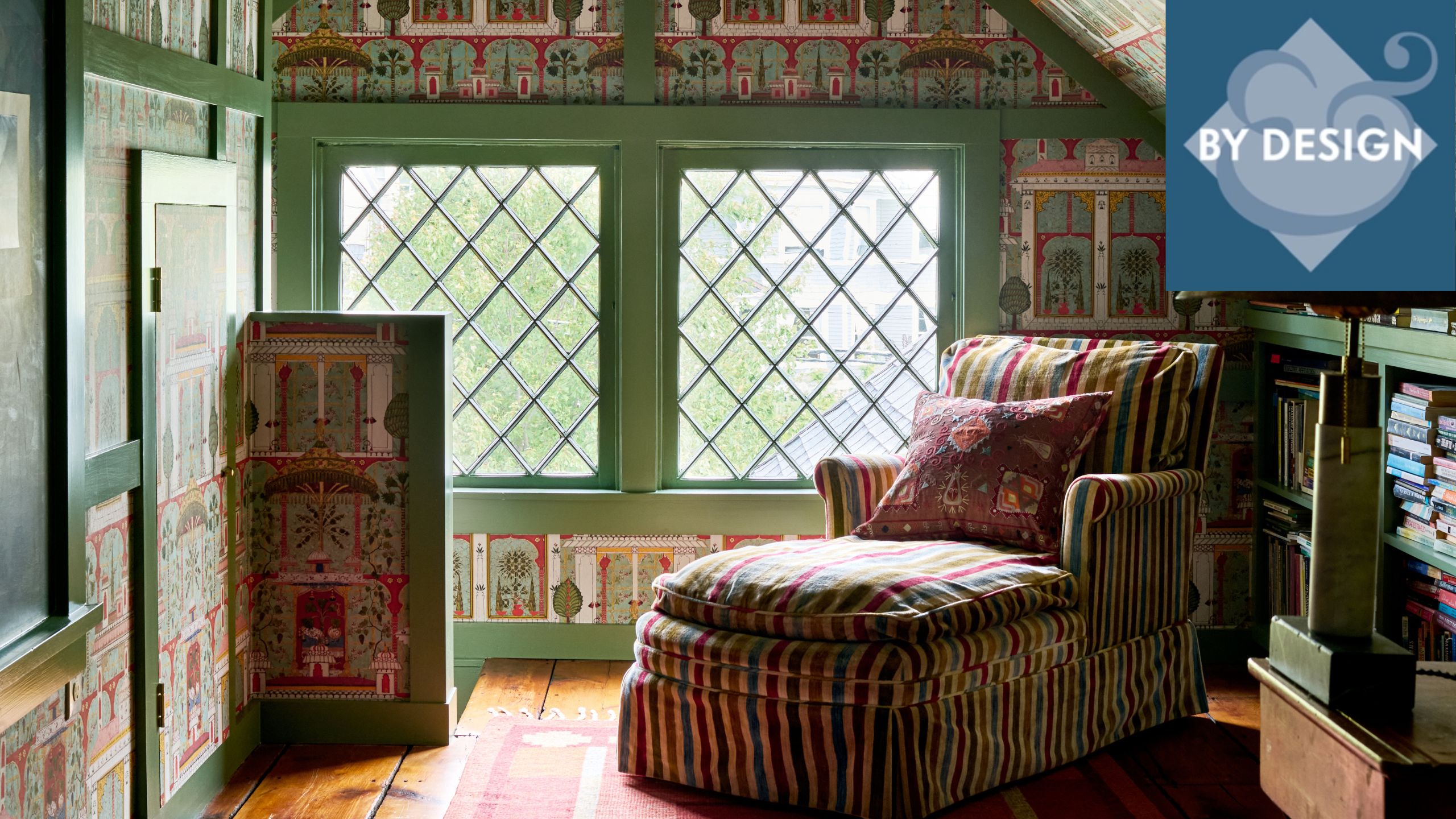 Colorful attic room with green painted trim, colorful wallpaper, striped chaise longue with patterned cushion, wooden floor with rug, bookshelf to the right.