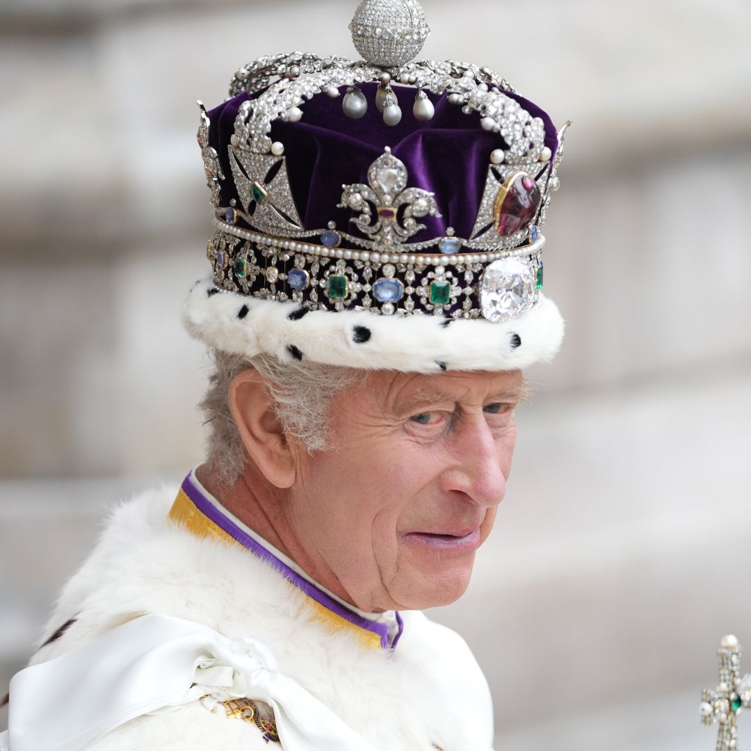 A headshot of King Charles wearing his coronation crown and robes in front of a marble column