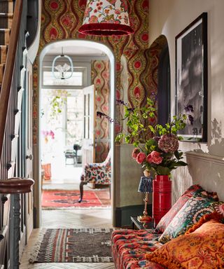 Vibrant, eclectic hallway design, patterned wallpaper, cozy seating area, textured patterned pendant light, wooden flooring, ornaments, vases on side table, artwork mounted above seating