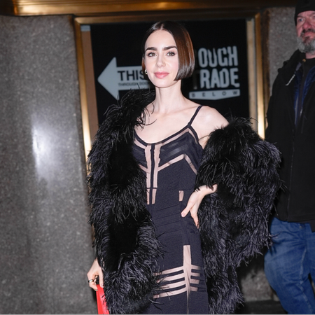 a paparazzi photo of a white woman with a short, dark bob. she is wearing a black dress with cutouts and a black feather coat