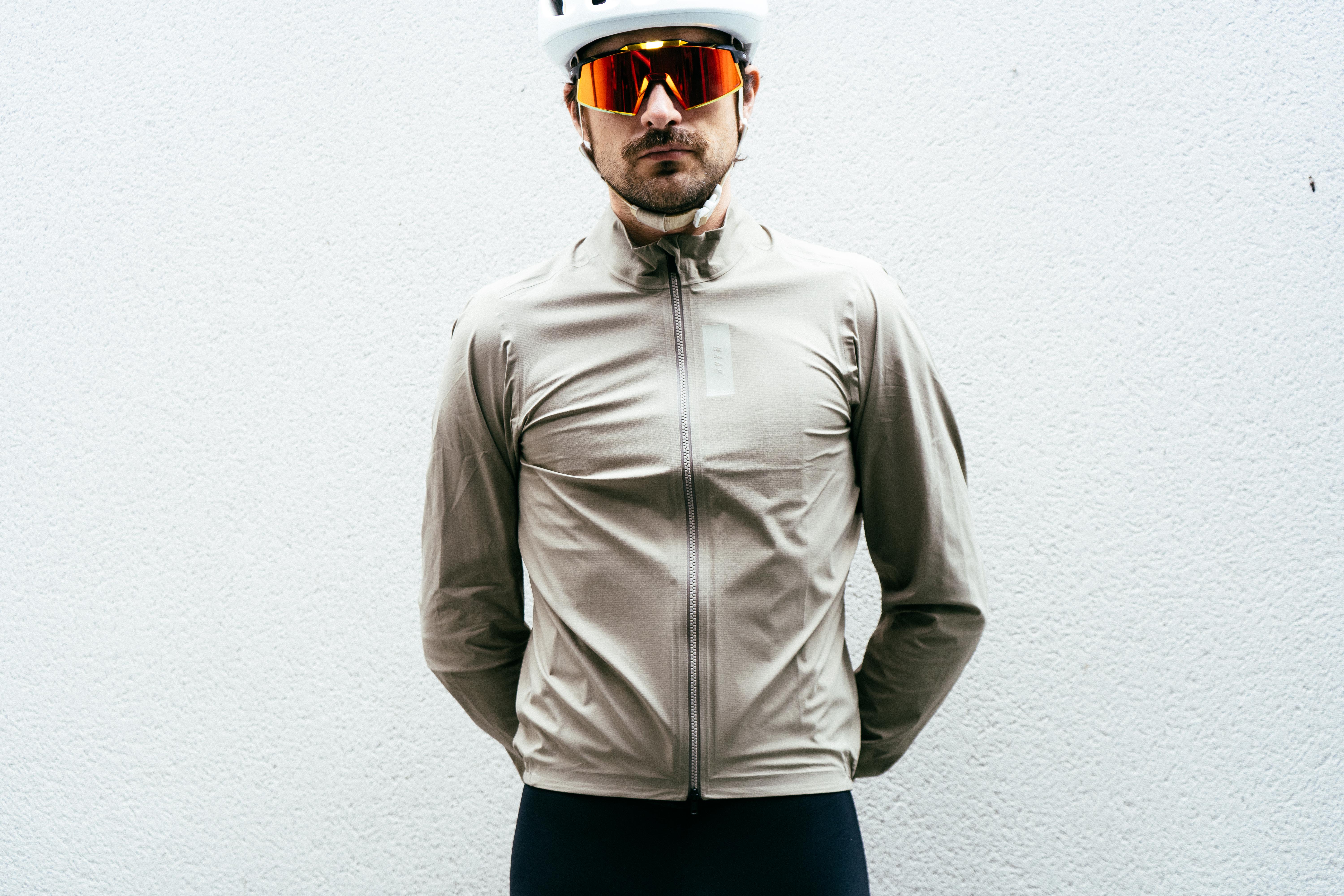 A man in an off-white waterproof cycling jacket stands against a white wall