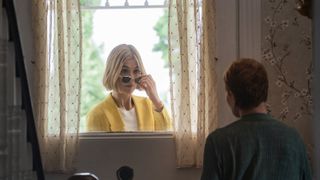 rosamund pike wearing a yellow sweater and dipping her sunglasses down while looking in a window at an older woman in i care a lot