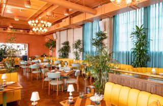 A warmly lit restaurant features light green and wooden retro-inspired chairs and yellow sofas, along with mid-century lighting and plants.