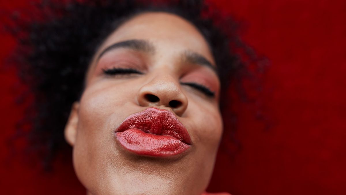 Smiling woman puckering red lips with eyes closed whilst standing against a red wall.