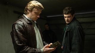 Kenneth Noye (Jack Lowden) in a brown leather jacket holding a gold bar next to Brian Robinson (Frankie Wilson) in The Gold