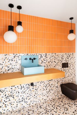 Bright orange small scale vertical metro tiles with terrazzo wall to floor tiling in modern bathroom complete with blue basin and black toilet