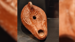 Ceramic oil lamp made out of orange-colored clay. There are two holes for the wick and the flame, and it is decorated with an incised menorah.