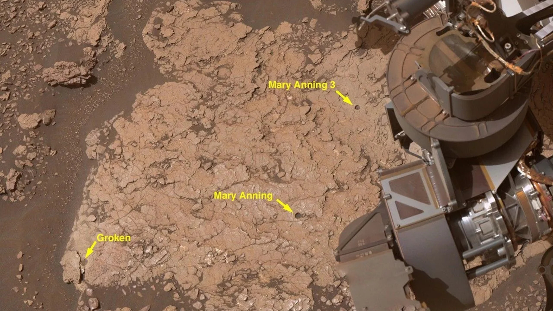 A close up of a reddish brown surface with a metal arm in the right top corner of the image. The image includes yellow arrows and labels of different sites of interest. 