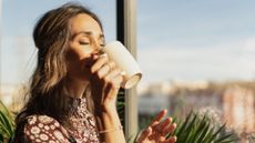 Woman drinking coffee from mug, standing at home in the sunshine after learning how to get rid of sensitive teeth with sensitive teeth treatments