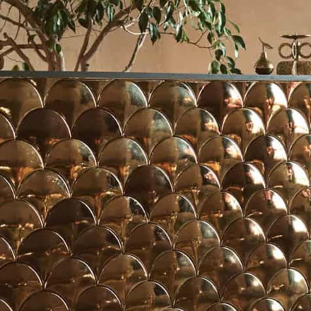 A close-up of a highly reflective metallic gold scallop-shaped tile splashback with a fish scale pattern, creating a warm, lustrous surface. A dark metal shelf above holds a small potted olive tree and decorative objects against a terracotta-toned wall.