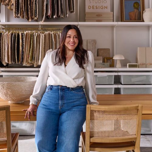 Image of a woman in jeans and a white top leaning on a wooden table with wooden chairs beside her.