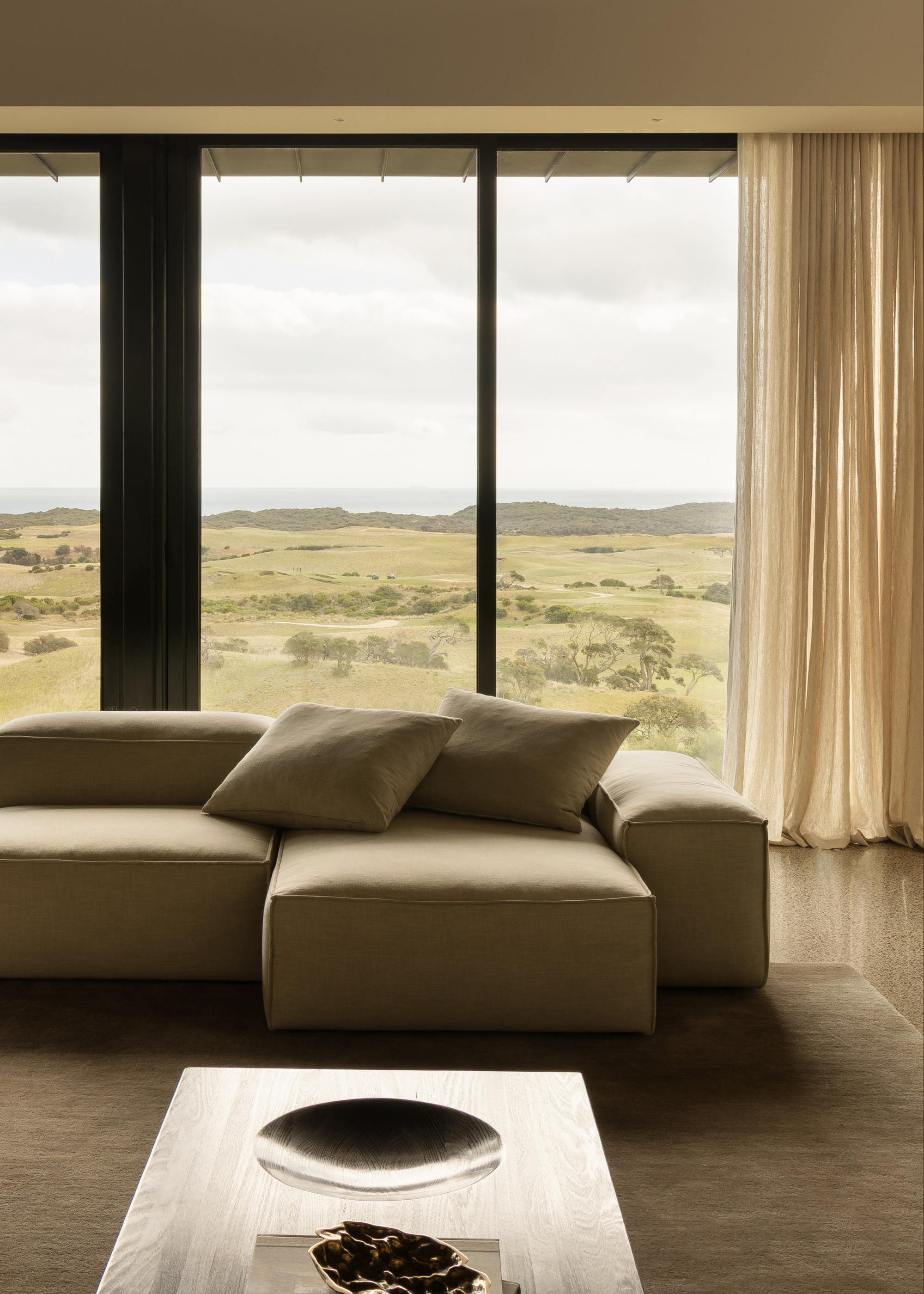 A living room with tall glass windows, sheer beige curtains, a beige sectional with pillows, a soft area rug, and a wood coffee table