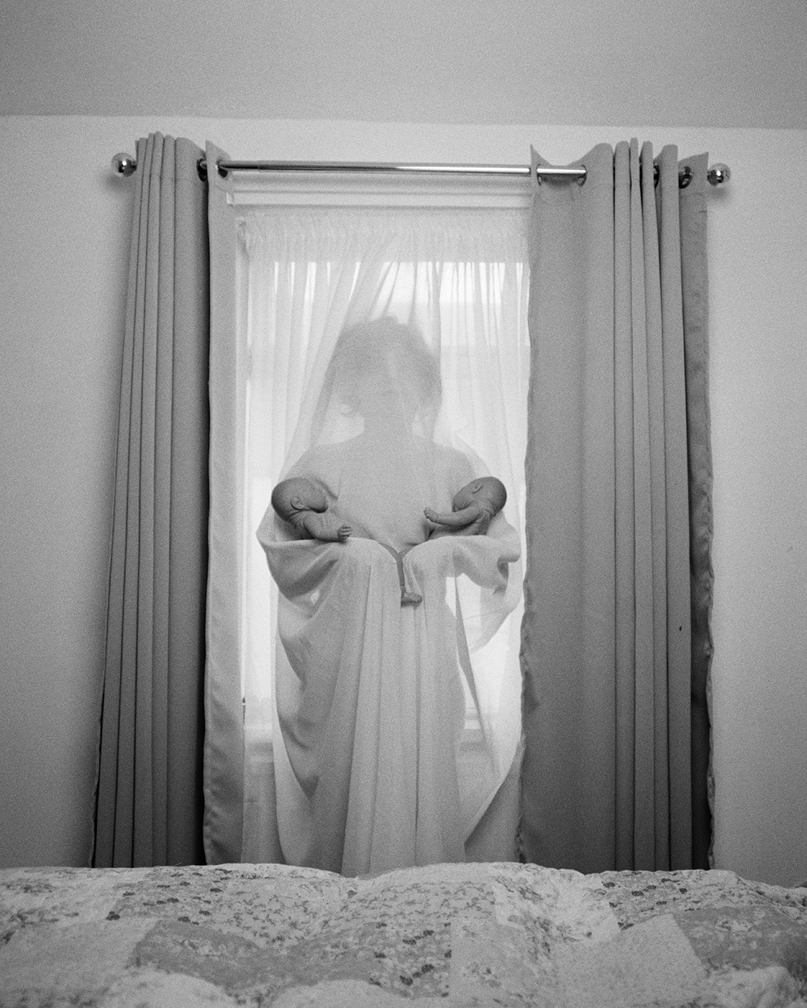 Black and white photograph of a woman behind a net curtain, holding two babies in each of her arms