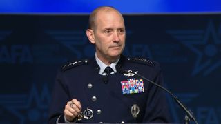 A man in a military uniform stands in front of a dark background. 