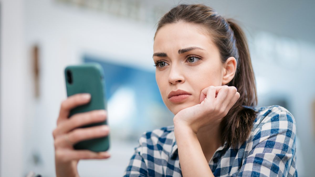 Close up of a depressed Caucasian young woman sitting at home, looking at her smartphone