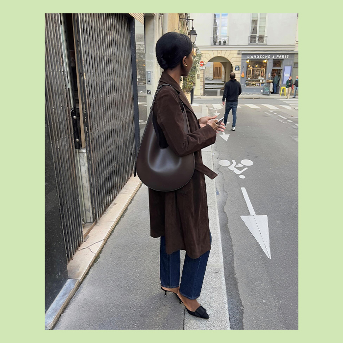 sylvie wears brown suede coat, jeans, black top, and deep brown leahter shoulder. bag