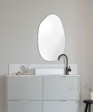 Gray bathroom with gray vanity unit, small white sink, black tap, oval asymmetrical mirror above, plant on black stool beside unit