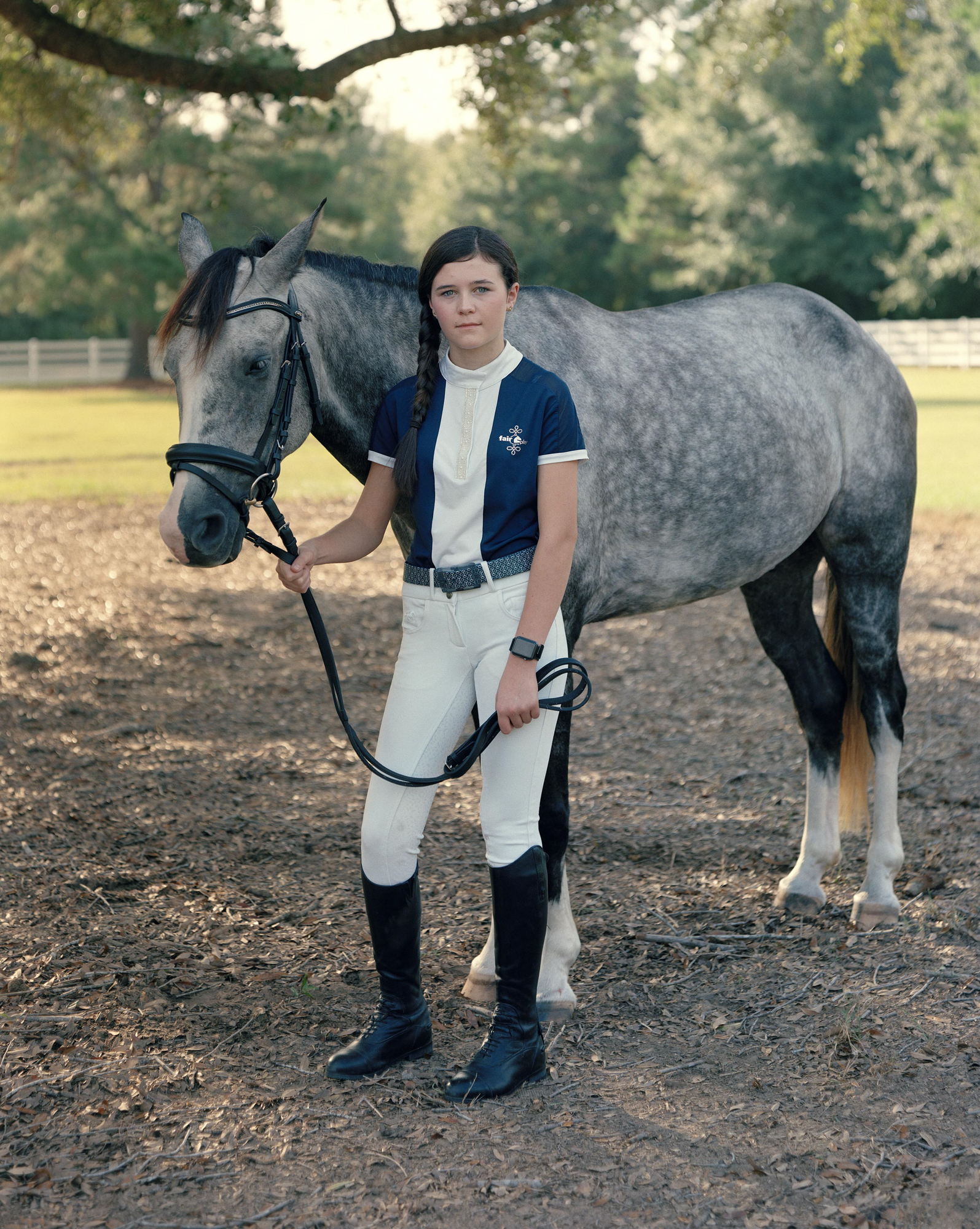 A girl in white riding breeches and tall black boots stands holding the reins of a dappled grey horse under a tree