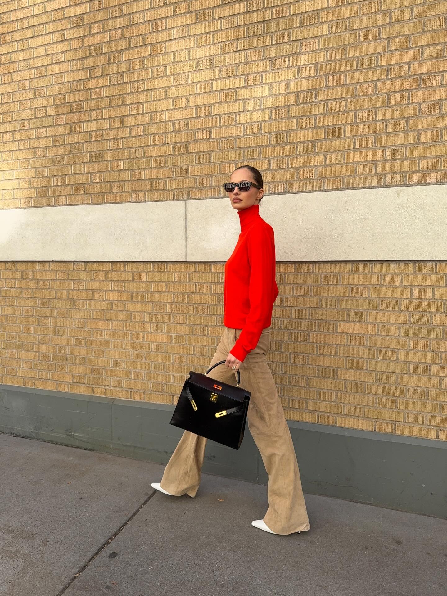 Pia Mance in New York City wearing a red turtleneck sweater with a pair of tan flare pants and a black Hermes Kelly bag.