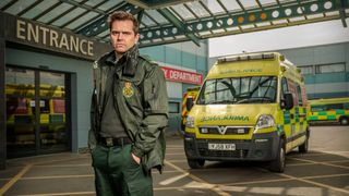 Michael Stevenson as paramedic Iain Dean looking seriously at the camera, standing in front of his ambulance and the entrance to Holby ED in Casualty. 