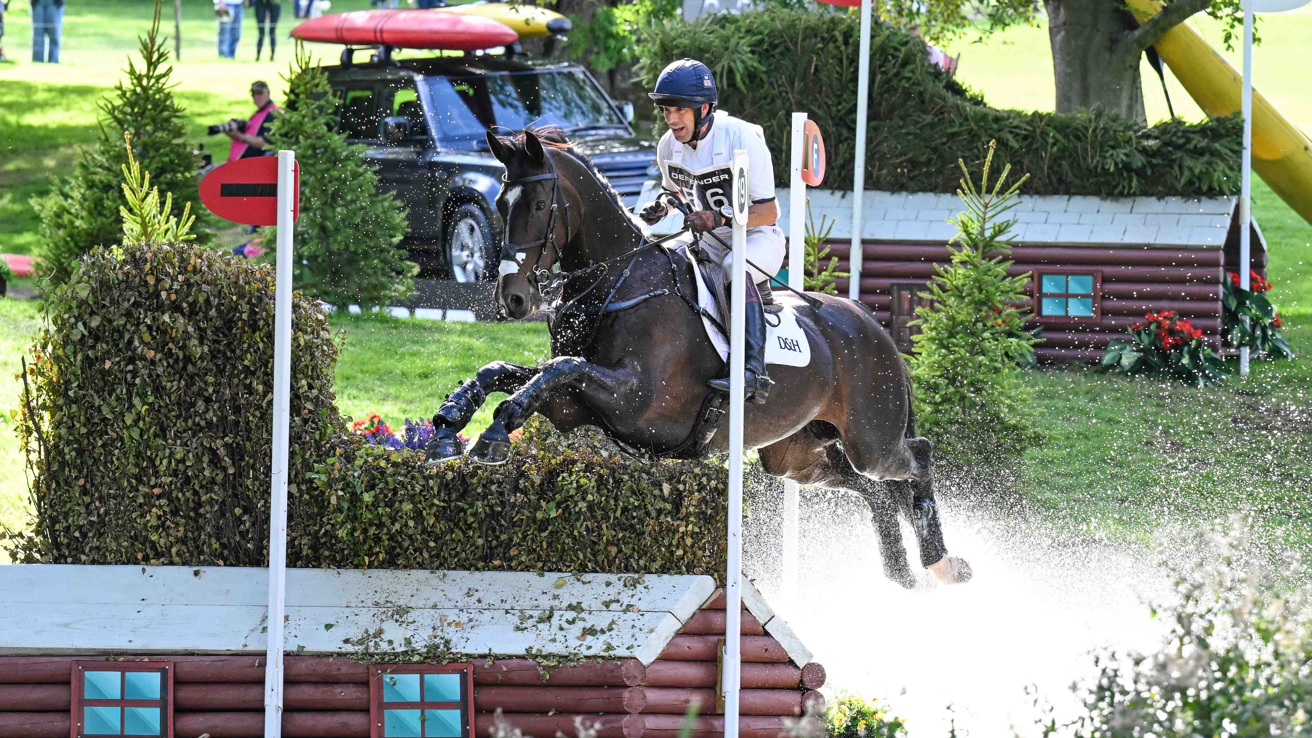 Harry Meade in white colours and a blue hat jumps a fence out of the water at the 2026 Defender Burghley Horse Trials riding Cavalier Crystal