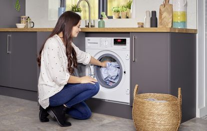 woman using the Beko Integrated 7kg 1600rpm Washing Machine with RecycledTub™ WTIK76151F, one of the best integrated washing machine picks, in her kitchen, putting clothes inside the machine