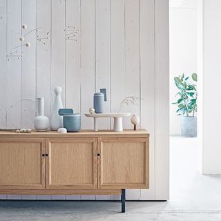 A room with white tongue and groove panelling and a wooden sideboard with curvaceous ceramic vessels on top