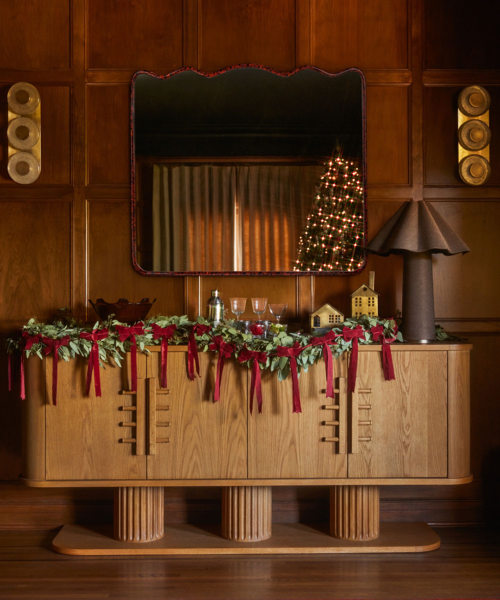 lulu and georgia christmas dining room shot with a wooden sculptural credenza styled with foliage garland, velvet bow ribbon garland, a mirror, brown scalloped lamp and small brass houses
