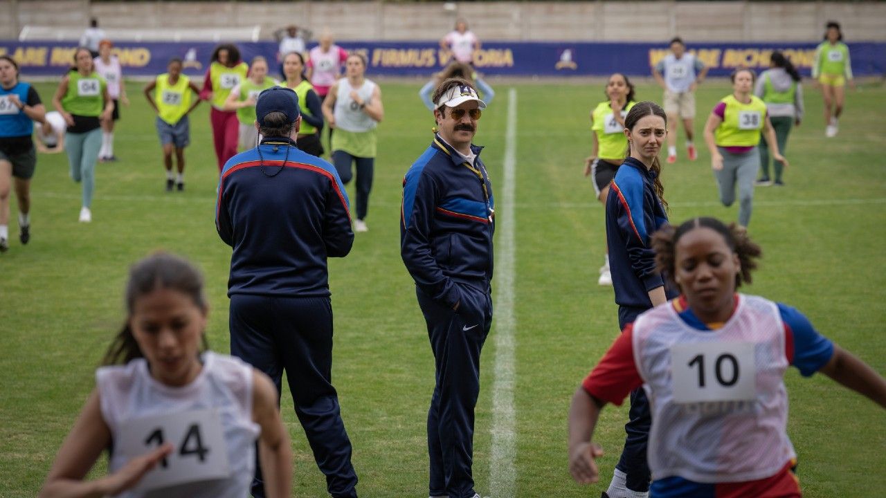 From left to right: Brendan Hunt looking forward, while Jason Sudeikis and Tanya Reynolds look back at women running. 