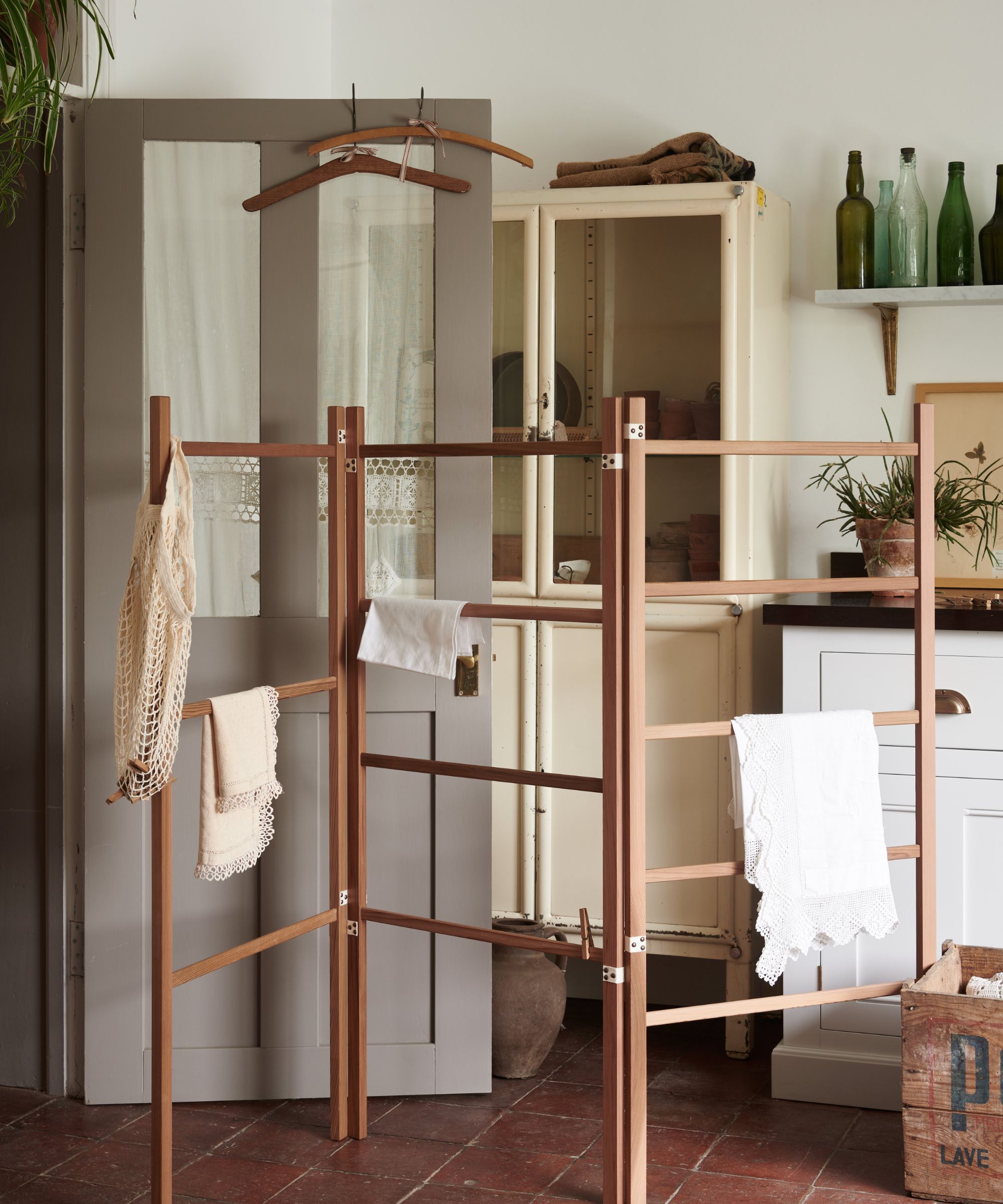 A rustic laundry room with a freestanding wooden clothes airer draped with lace linens, a vintage cream glazed cabinet, terracotta tile floor and green glass bottles on a shelf.