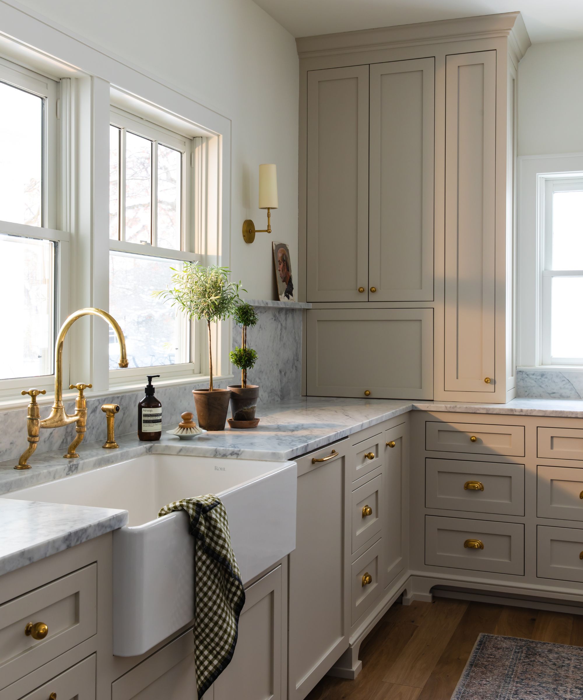 A modern American kitchen with stone-colored cabinetry, a white butler's sink in front of two windows, white walls, brass fittings, and marble countertops.
