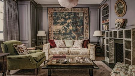 a mauve living room with a large vintage tapestry, wall paneling, a unique square fireplace, cream velvet sofa and green armchair