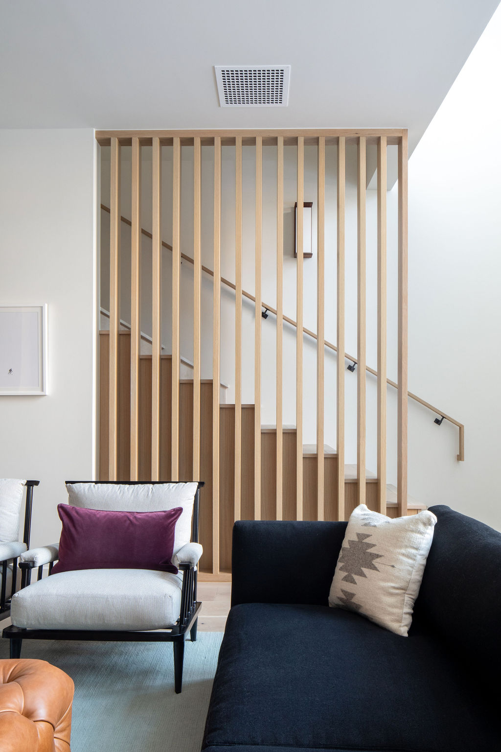 Interior with blue oversized couch, white and black armchairs, cushions, and a central leather ottoman, set in front of a wooden staircase.