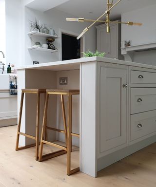 An angled shot of a white kitchen island wih two brushed brass bar stools underneath it and a gold chandelier and white shelves and walls above it