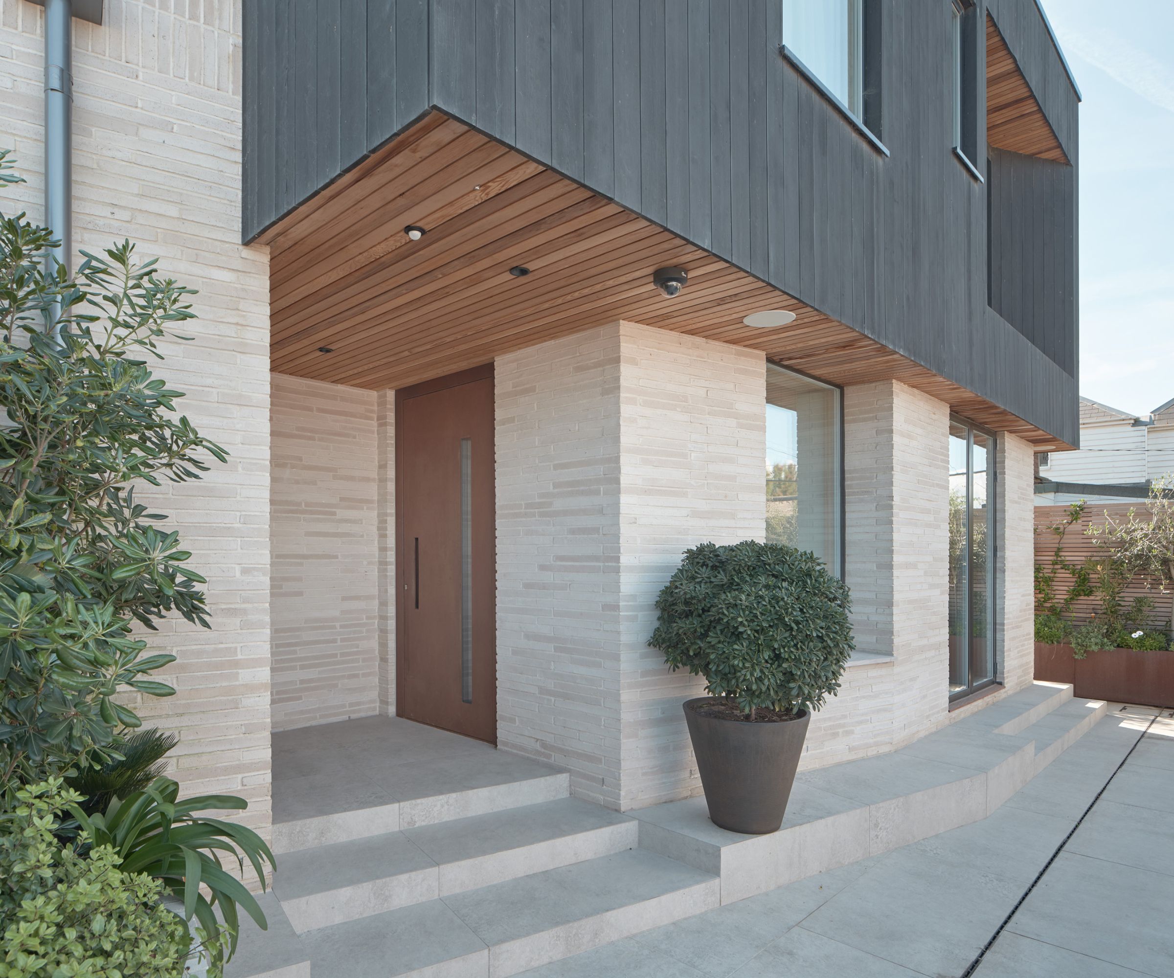 A modern home entrance featuring a wide brown door, light stone walls, a wood-panelled ceiling, and large potted plants flanking the steps.