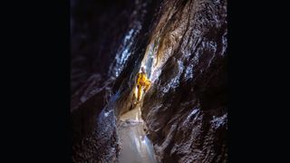 A man wearing a hard hat and a yellow jumpsuit looks up the wall of a narrow dark cave.