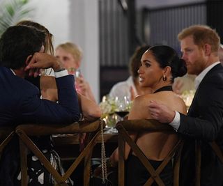 Meghan Markle sits at a dinner table with Prince Harry wearing a backless black St Agni dress and vintage earrings