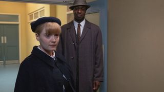 Rosalind (Natalie Quarry) in uniform stands in a lobby with Cyril (Zephryn Taitte) in a dark suit and hat in Call the Midwife.