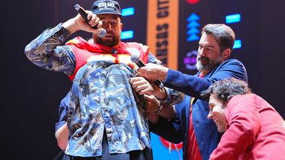 Travis Kelce puts on a jersey as he stands onstage with Rob Riggle and Paul Rudd during 2024 Big Slick Celebrity Weekend on June 01, 2024 in Kansas City, Missouri.