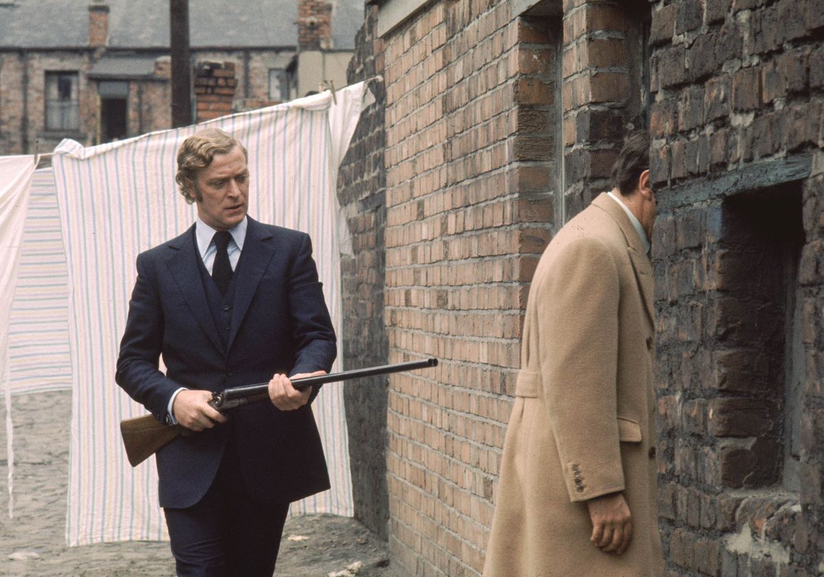 Michael Caine points a shotgun at the back of a man entering a house