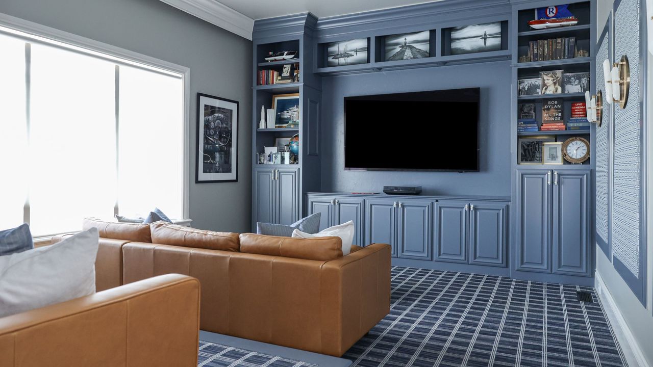 Small theater room ideas are so chic. Here is a small blue theater room with a bookcase around the black rectangular TV, a light brown leather couch facing it, and blue and white grid carpet on the floor