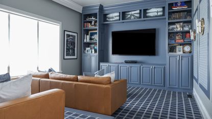 Small theater room ideas are so chic. Here is a small blue theater room with a bookcase around the black rectangular TV, a light brown leather couch facing it, and blue and white grid carpet on the floor