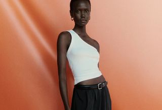 Woman wearing white one-shoulder tank top, and black flowy skirt with leather belt against orange studio backdrop.