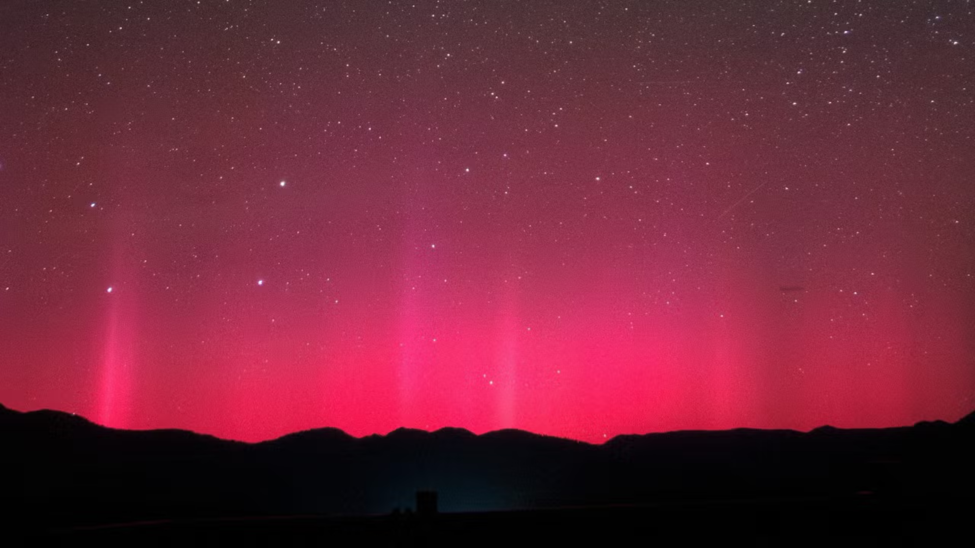 The silhouette of what looks like mountains on the bottom of the screen with bright pink and red lights in the sky above. 