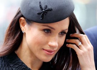 Meghan Markle wearing a black beret and holding up her hand to show her diamond engagement ring