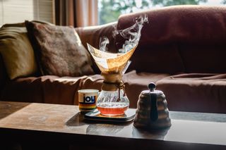 An image of a Chemex pour over coffee brewer, set on a coffee table between a colorful mug and a metal kettle